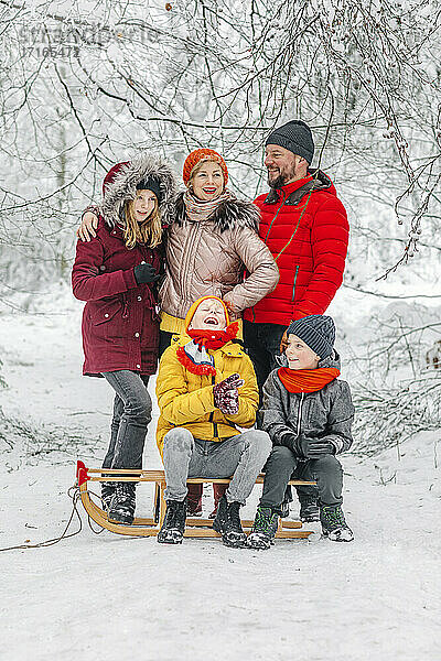 Eltern stehen bei Kindern  die im Winter im Wald auf einem Schlitten sitzen