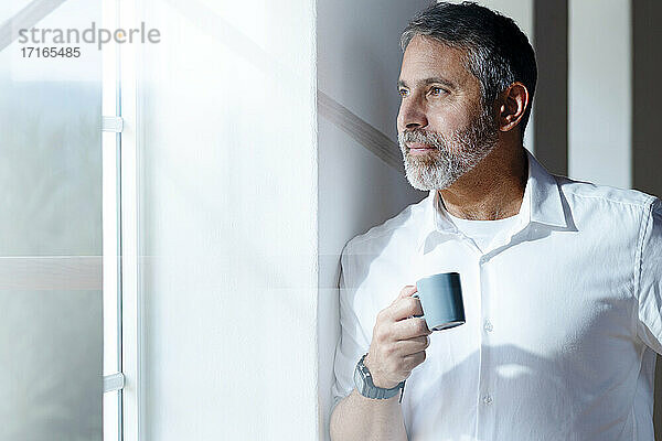 Älterer Geschäftsmann mit Kaffeetasse  der durch das Fenster schaut  während er zu Hause steht