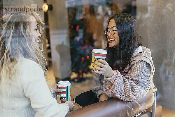 Young female friends having coffee while sitting in cafe seen through window glass