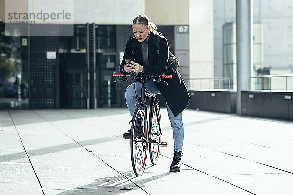 Young woman using mobile phone on bicycle outside building