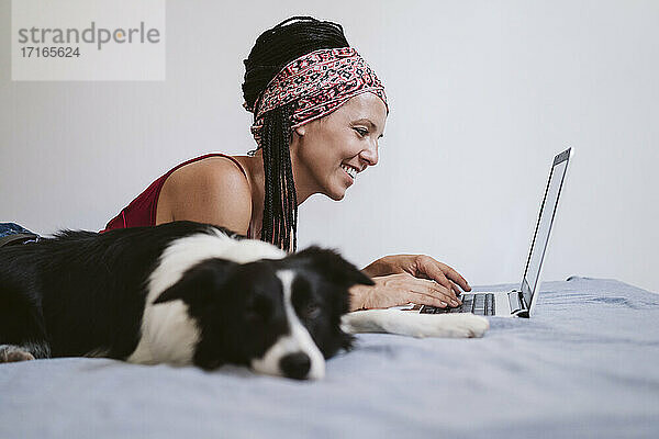 Lächelnde Frau  die einen Laptop benutzt  während sie zu Hause auf dem Bett bei ihrem Haustier liegt