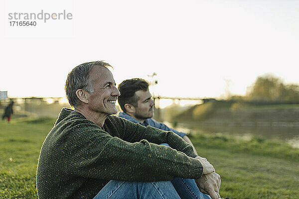 Smiling father spending leisure time with son on grass at riverbank