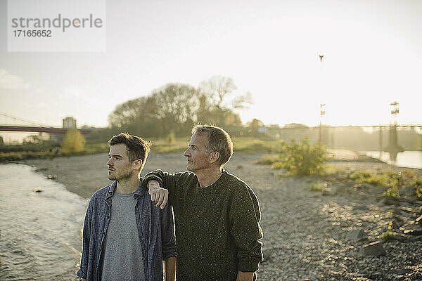 Father and son looking away while standing on riverbank at evening