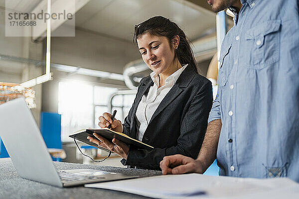 Smiling businesswoman writing in book while standing by colleague at industry