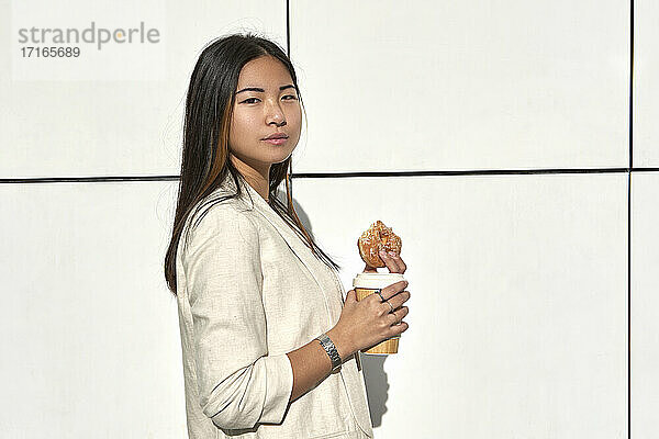 Confident young woman standing with reusable cup and doughnut against wall