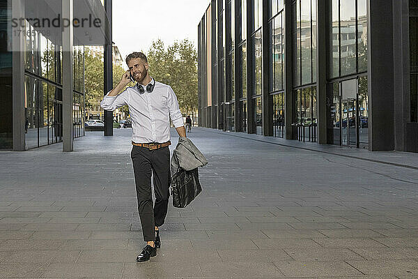 Smiling businessman with briefcase talking on mobile phone while walking on footpath