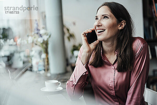 Lächelnde Unternehmerin  die mit ihrem Smartphone spricht und im Café nach oben schaut