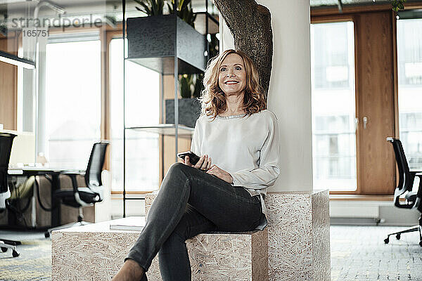 Lächelnde Geschäftsfrau mit Smartphone  die auf einer Stützmauer im Kreativbüro sitzt
