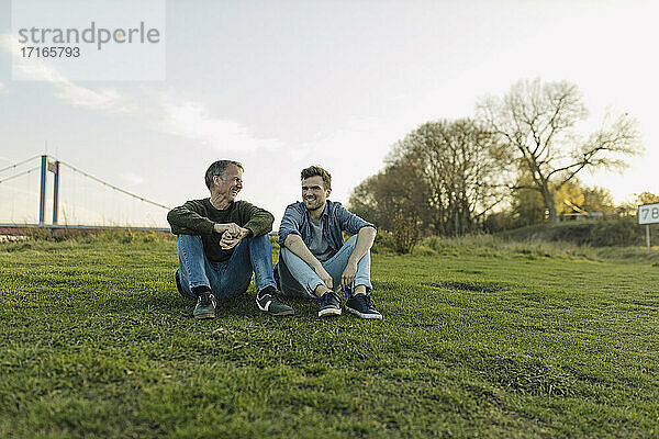 Happy father and son talking with each other while sitting on grass against sky