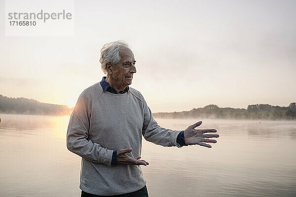 Älterer Mann gestikuliert  während er bei Sonnenaufgang am See steht