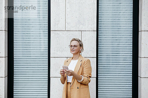 Ältere Geschäftsfrau mit Smartphone  die stehend gegen eine Wand schaut