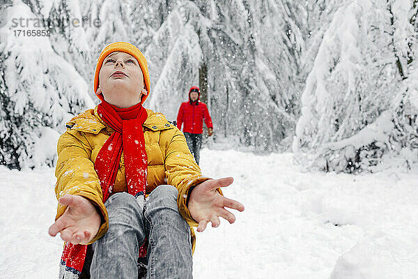 Junge schaut nach oben  während er mit einem Mann im Hintergrund im Wald sitzt  während es schneit