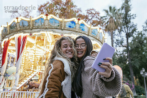 Multi-ethnic female friends talking selfie through mobile phone against illuminated carousel