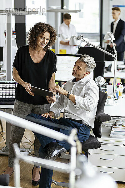 Geschäftsmann und Frau arbeiten an einem digitalen Tablet mit Kollegen im Hintergrund in einem Großraumbüro