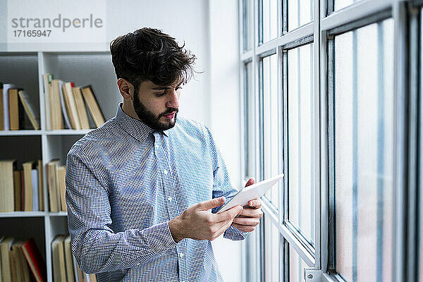 Junger Geschäftsmann benutzt digitales Tablet am Fenster in einem kreativen Büro