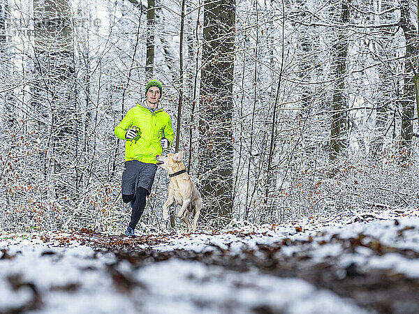 Junger männlicher Sportler joggt mit Labrador Retriever im Winter im Wald