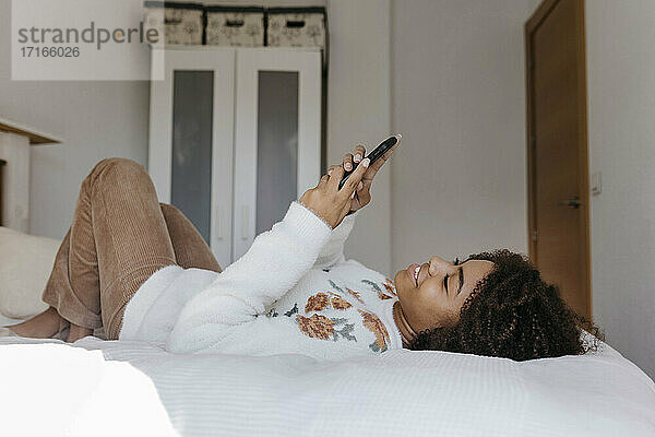Junge Frau benutzt ein Mobiltelefon  während sie sich zu Hause auf dem Bett ausruht