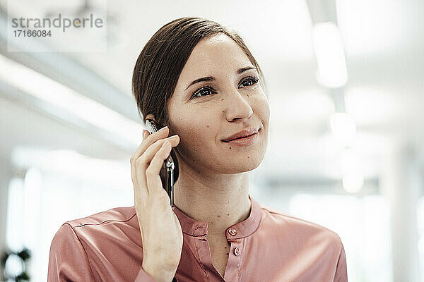 Lächelnde Geschäftsfrau  die wegschaut  während sie mit einem Smartphone im Büro spricht