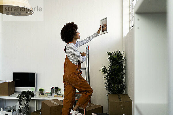 Afro-Frau  die einen Bilderrahmen an die Wand hängt  während sie auf eine Leiter in ihrer neuen Wohnung steigt