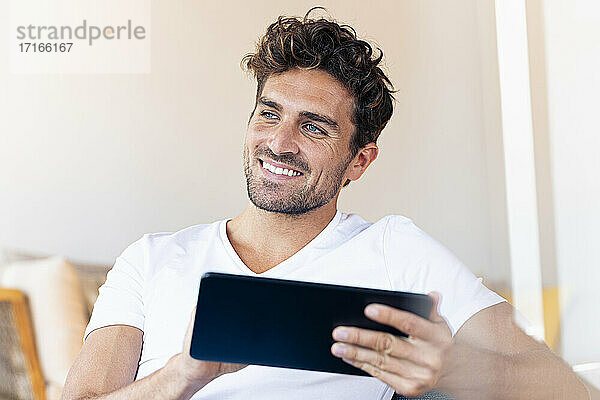 Nachdenklicher Mann mit digitalem Tablet  der wegschaut  während er zu Hause sitzt