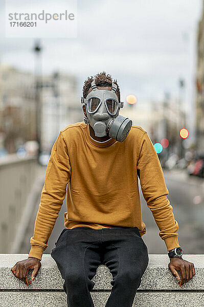 Young man wearing gray gas mask sitting on retaining wall
