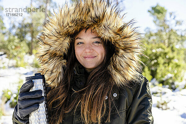 Lächelnde Frau mit Pelzkapuzen-Wintermantel  die eine Flasche hält  während sie im Wald steht