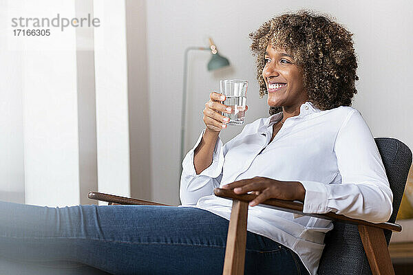 Lächelnde Frau  die ein Wasserglas hält  während sie zu Hause auf einem Sessel sitzt