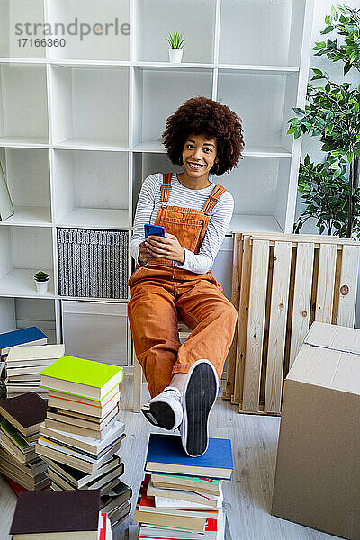 Lächelnde Afro-Frau  die ihr Handy in der Hand hält  während sie an einem Bücherregal in ihrem neuen Zuhause sitzt
