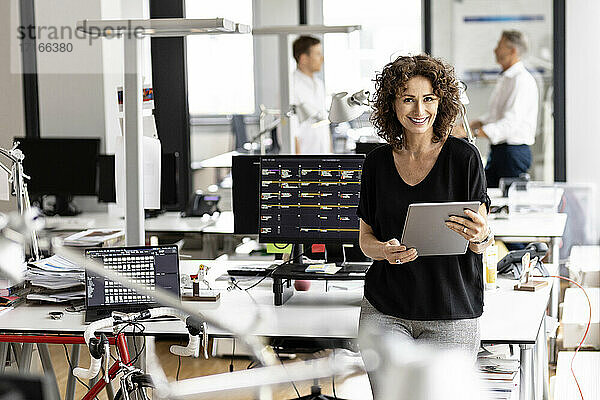 Lächelnde Geschäftsfrau  die ein digitales Tablet hält  während sie mit einem Kollegen im Hintergrund in einem Großraumbüro steht