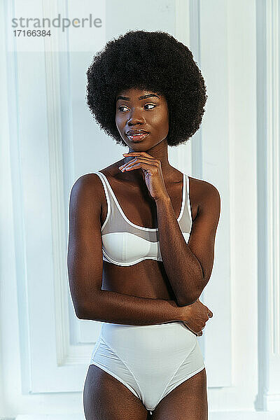 Afro junges Model mit Hand am Kinn in weißen Dessous schaut weg gegen Wand