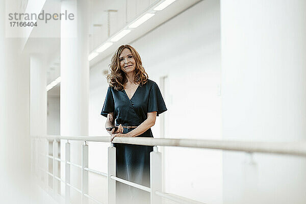 Lächelnde Geschäftsfrau mit Smartphone am Geländer lehnend im Korridor