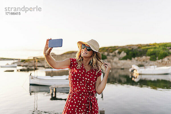 Lächelnde Frau mit Hut  die ein Selfie mit ihrem Mobiltelefon macht  während sie gegen den Himmel steht