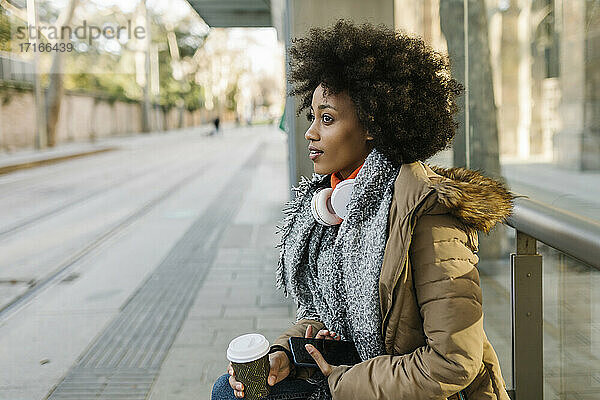 Afro-Frau mit Smartphone und Einweg-Kaffeebecher nachdenklich am Busbahnhof im Winter