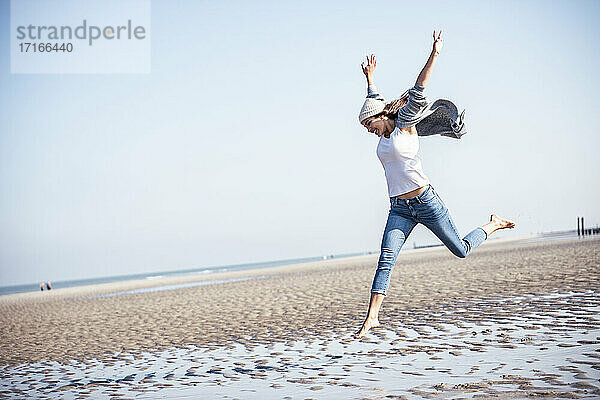 Fröhliche junge Frau mit erhobenen Armen  die über nassen Sand am Strand springt