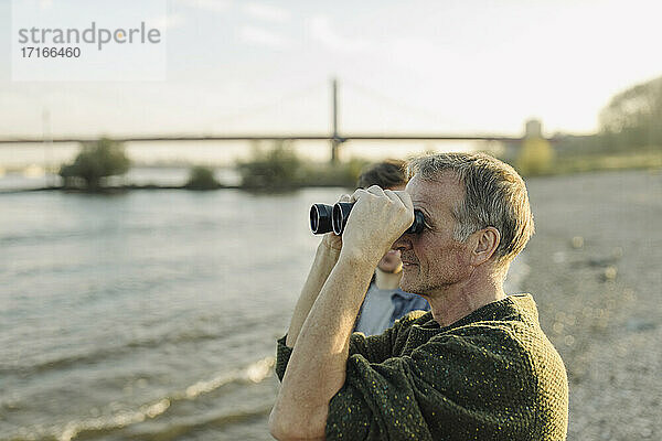 Father looking through binoculars standing with son on riverbank at evening