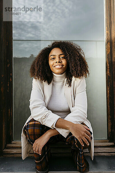 Lächelnde afroamerikanische junge Frau in weißem Mantel auf dem Fensterbrett sitzend