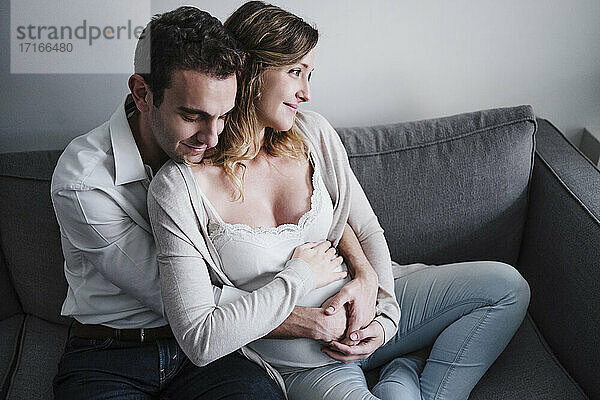 Mann mit geschlossenen Augen  der seine schwangere Frau umarmt  während er zu Hause auf dem Sofa sitzt