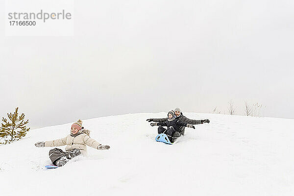 Verspielter Vater mit Kindern beim Schlittenfahren auf einem verschneiten Hügel gegen den Himmel im Urlaub