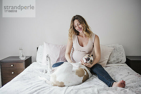 Lächelnde schwangere Frau spielt mit Hund auf Bett zu Hause