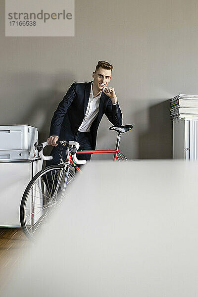 Selbstbewusster Geschäftsmann mit Hand am Kinn  der sich auf ein Fahrrad stützt  während er im Büro steht