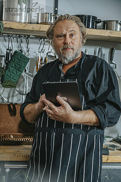 Mature chef wearing apron using digital tablet while standing in kitchen