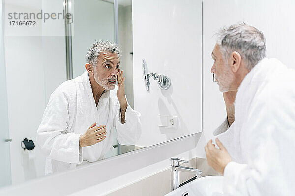 Surprised man wearing bathrobe looking at mirror in hotel bathroom