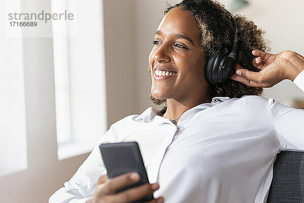 Glückliche Frau mit Kopfhörern  die ein Mobiltelefon benutzt  während sie zu Hause sitzt