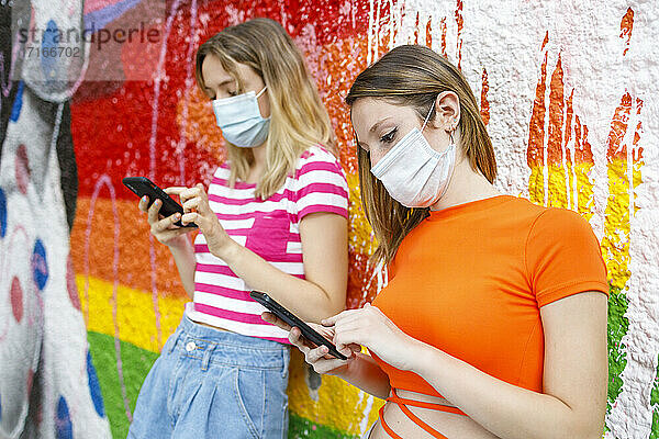 Freundinnen benutzen ein Smartphone  während sie sich während COVID-19 an eine bemalte Wand lehnen