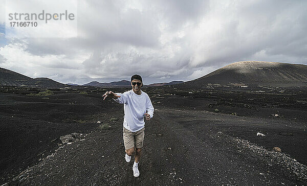 Lächelnder männlicher Tourist  der auf schwarzer Erde am Vulkan El Cuervo  Lanzarote  Spanien  läuft und gestikuliert