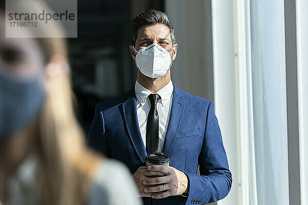 Geschäftsmann mit Gesichtsschutzmaske und Kaffee in der Hand im Büro stehend