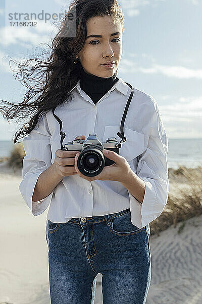Beautiful young woman holding camera while standing at beach against sky