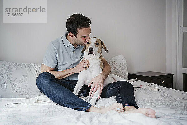 Mittlerer erwachsener Mann küsst Hund  während er zu Hause auf dem Bett an der Wand sitzt