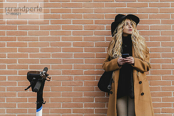 Woman in coat using smart phone while standing against brick wall