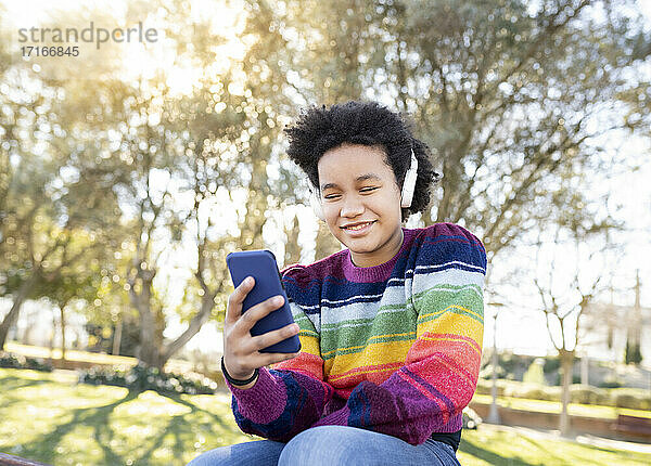 Lächelndes Mädchen mit Kopfhörern  das ein Mobiltelefon benutzt  während es im Park sitzt